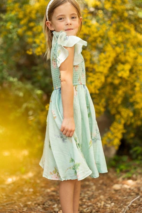 A young girl with light brown hair, wearing the Mini Hattie Dress in Roselyn Blue by Ivy City Co—a pale blue floral dress featuring a smocked bodice and crafted from a soft cotton-linen blend—stands outdoors on a dirt path with yellow flowers and greenery in the background.