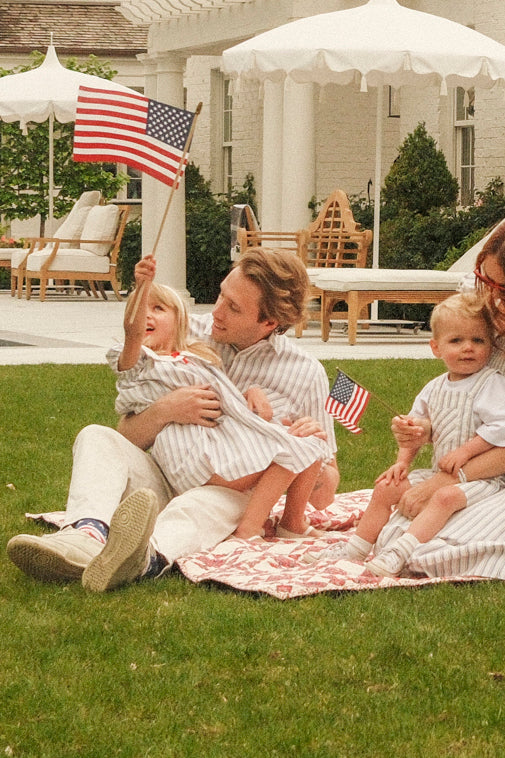A man in the Ivy City Co Men's James Shirt in Heritage Stripe sits on a blanket with three kids in matching striped outfits; two hold small American flags. Outdoor seating and umbrellas are seen in the background.