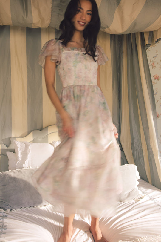 Wearing the Ivy City Co Rhiannon Dress in Painted Bloom, a woman with long dark hair stands smiling on a bed in a softly lit, elegant bedroom with striped curtains and plush pillows.