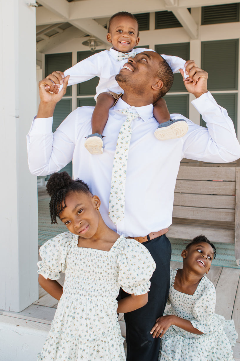 A smiling man in a white shirt and Ivy City Co Ties in Madeline Blue Floral carries a young boy on his shoulders, while two girls in coordinating floral dresses stand happily by his side—each perfectly styled in matching Ivy City Co family outfits.