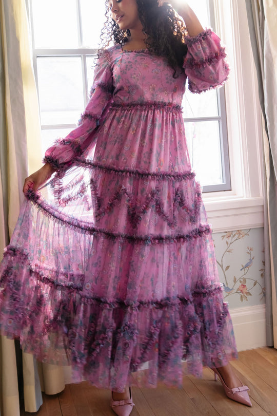A woman stands indoors by a window, holding out the skirt of her long, sheer, ruffled floral Catherine Dress in Climbing Vine Bloom by Ivy City Co. Sunlight filters behind her as she wears matching pink heels, highlighting the fresh elegance of this spring piece from Ivy City Co’s Climbing Vine Bloom collection.