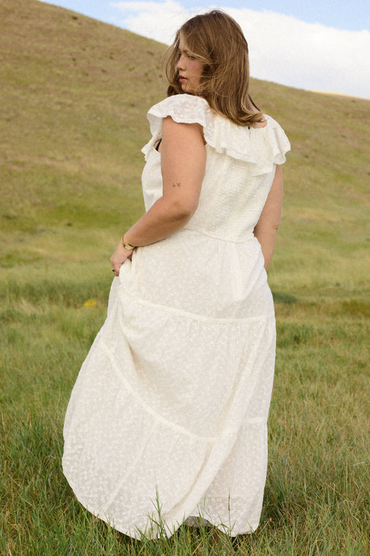 Wearing the Alaina Dress in Ivory by Ivy City Co, a woman stands in a grassy field with her back turned and head glancing to the side. Behind her, rolling hills and a partly cloudy sky complete the serene backdrop.