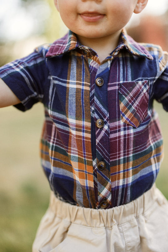 A young child wearing the Ivy City Co Baby Boy James Romper in Midnight Plaid stands outdoors, arms slightly raised, with their face partially visible and smiling.