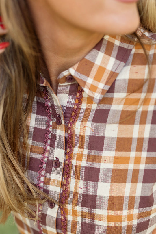A close-up of a person wearing the Ivy City Co Bowie Long Sleeve Dress in Spiced Plaid, featuring brown, orange, and cream plaid cotton fabric with buttons and decorative stitching. The person’s light brown hair is partially visible.