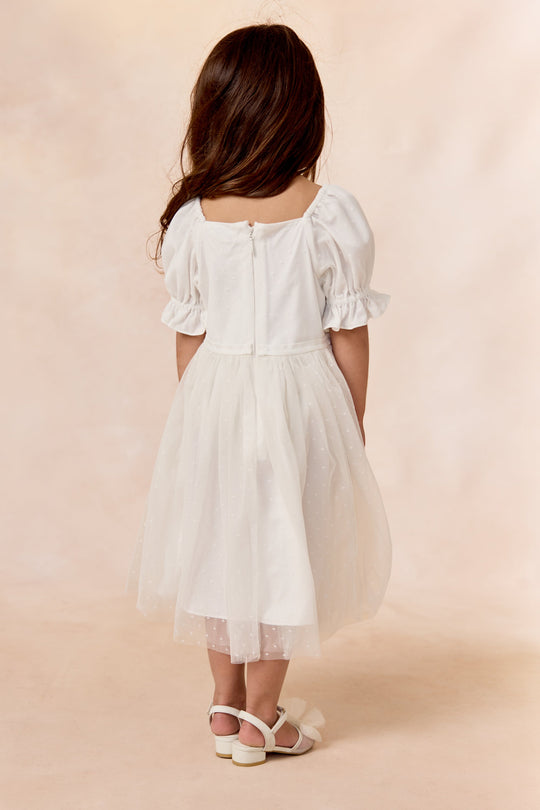 A young girl with long brown hair, wearing the Ivy City Co Mini Ballerina Dress in White—featuring puffed sleeves and a delicate tulle skirt—and white sandals, stands facing away from the camera against a soft beige background.