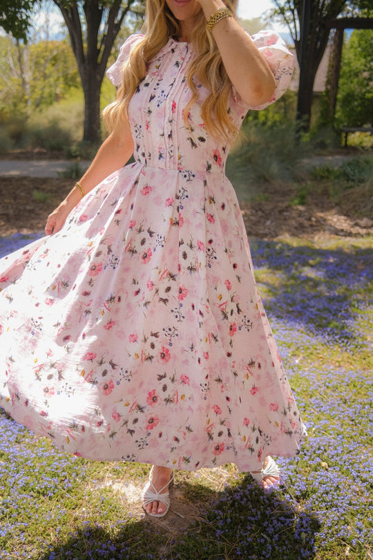 A woman wearing the Ivy City Co Sophia Dress in Roselyn Pink twirls outdoors on a sunny day, standing on a patch of purple flowers with trees in the background. Her face is partially obscured by her hand and hair.