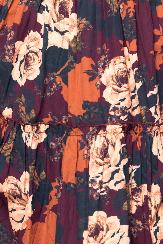 Close-up of the fabric from the Madeline Dress in Equinox Rose by Ivy City Co—a floral midi dress adorned with large white flowers and green leaves against a background of rust orange and deep burgundy. The gathered fabric, accentuated by a visible horizontal seam, gives this style an elegant look that is also maternity friendly.