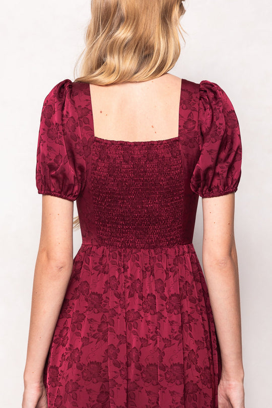 A woman with long blonde hair is seen from behind wearing the Ivy City Co Amira Dress in Red, a burgundy jacquard maxi dress adorned with a floral pattern, puff sleeves, and a smocked back. The image emphasizes the gathered fabric and square neckline of the dress.