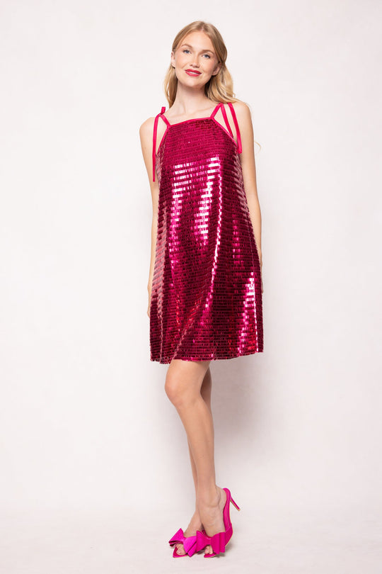 A woman stands and smiles in the Ivy City Co Zelda Dress, featuring fuchsia shingle sequins, a sleeveless design, and playful shoulder ties. She completes the look with matching pink high-heeled shoes adorned with bows. Set against a white background.