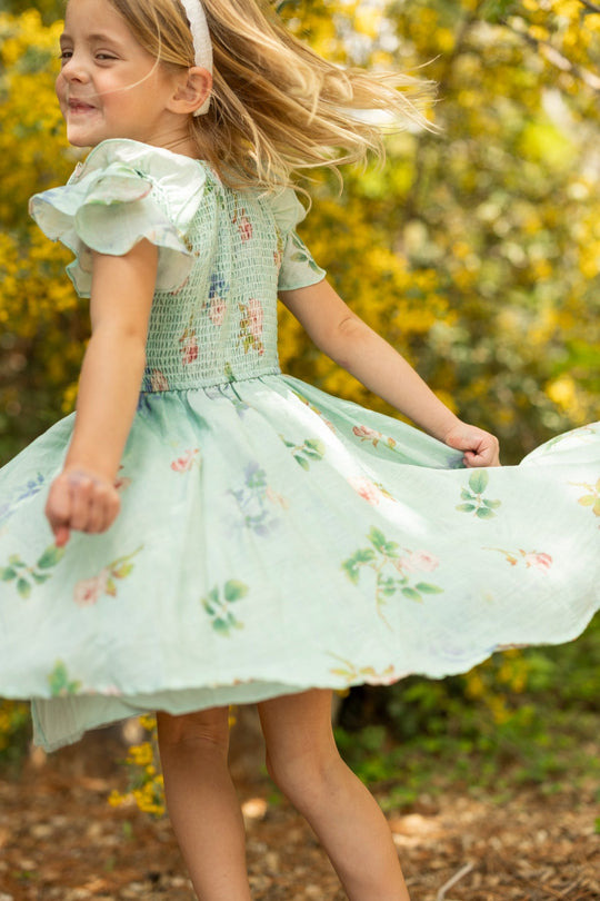 A young girl in an Ivy City Co Mini Hattie Dress in Roselyn Blue and headband twirls joyfully outdoors, surrounded by sunlight and yellow flowers.