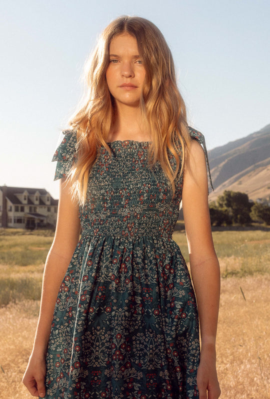 tween girl in a floral dress standing in a field with mountains in the background