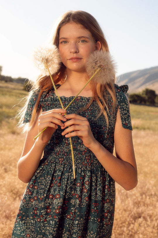 Young girl in a floral dress holding a dandelion in a field with mountains in the background