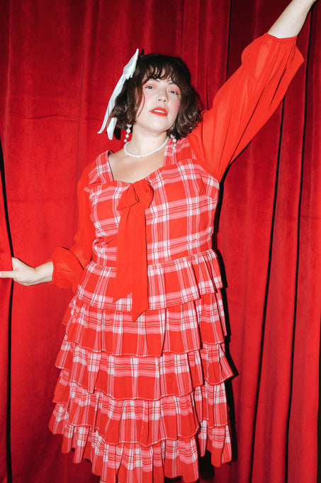 A person confidently poses in front of matching red curtains, wearing the Ivy City Co Taylor Dress in Red Plaid—a tiered dress with a large bow, pearl necklace, and white hair accessory—showcasing this bold women’s look with flair.