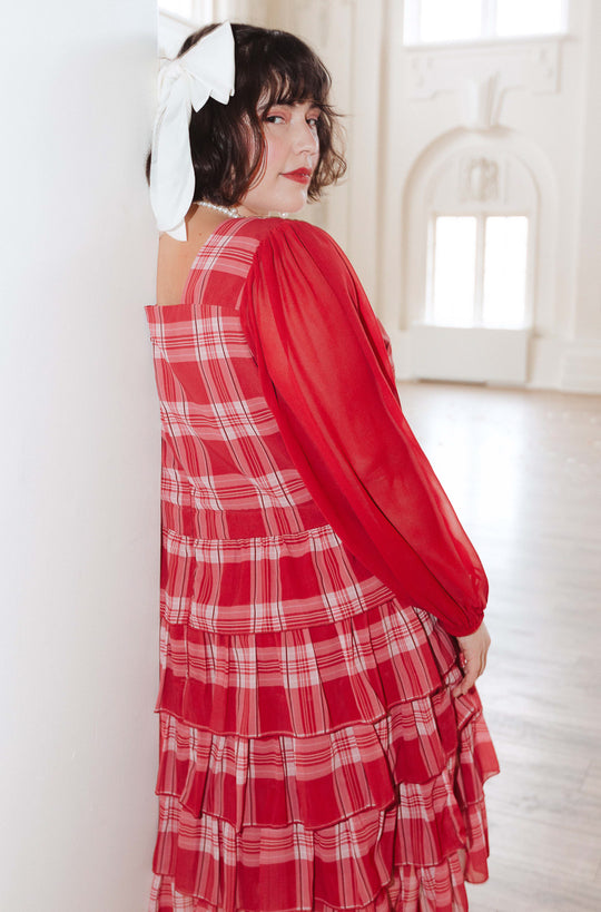 A person with short dark hair and a large white bow poses in a bright room, modeling the Taylor Dress in Red Plaid by Ivy City Co, featuring ruffled layers and sheer long sleeves, while looking over their shoulder at the camera.