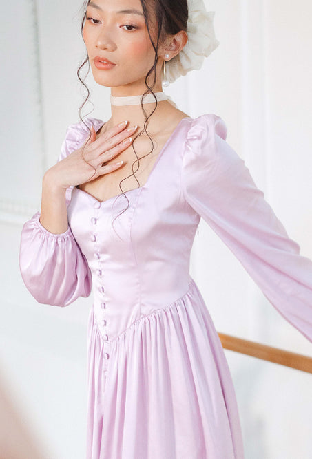 A woman poses gracefully indoors wearing the Ivy City Co Valentina Dress in Lavender, featuring satin fabric, long sleeves, a lace-up corset back, vintage-style buttons, and paired with a white choker and hair accessory, her curls styled loosely.