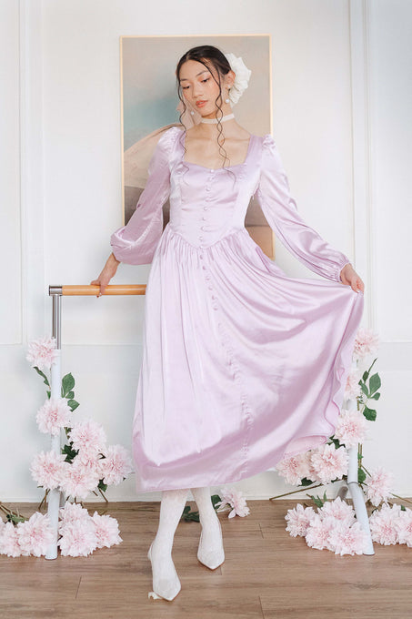 A woman models the Ivy City Co Valentina Dress in Lavender, featuring shiny satin, buttons, and a lace-up corset back. She poses indoors with white shoes and a hair accessory, surrounded by pink flowers and pastel art.