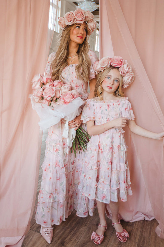 A woman and young girl wear matching Ivy City Co Mini Winnie Dresses in Pink Floral, featuring smocked bodices. Standing before pink curtains with large rose headpieces, the woman holds a bouquet as they exude soft, elegant charm.