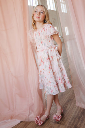 A young blonde girl stands indoors near sheer pink curtains on a wooden floor, wearing the Ivy City Co Mini Winnie Dress in Pink Floral with pink sandals. She poses gracefully, looking slightly upward with a gentle expression.