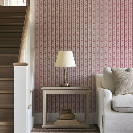A beige sofa sits beside a small wooden table with a lamp and woven baskets, next to a staircase. The wall behind showcases the Felicity Floral Wallpaper by Loomwell Home Goods, featuring custom-printed pink and red vertical stripes with elegant floral designs.