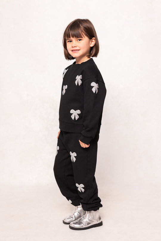 A young child with straight brown hair wears Ivy City Co’s Mini Micah Jogger Sweatpants in Black along with a coordinating sweatshirt, both adorned with silver bow designs reminiscent of rhinestone bow joggers. The outfit is paired with silver shoes and sparkly socks. The child stands sideways and smiles against a plain light background.