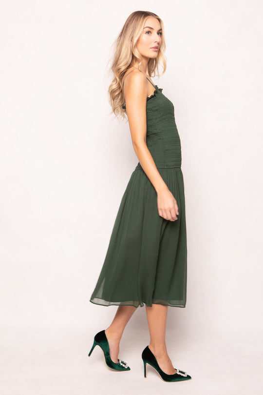 A woman with long blonde hair stands sideways, wearing the Ivy City Co Kimberly Dress in Green—a sleeveless, maternity-friendly midi dress with a smocked bodice and flowy skirt—paired with dark green high heels. She poses against a plain light background.