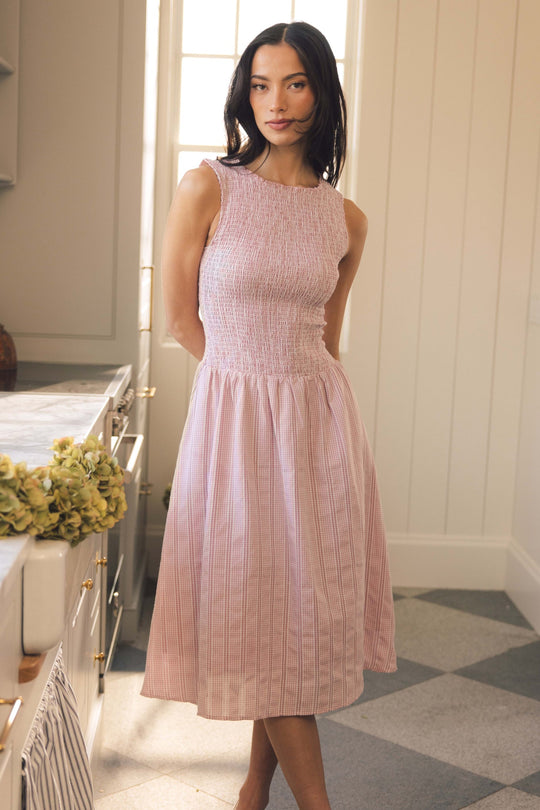 A woman wearing the Karla Dress in Pink Striped Gingham by Ivy City Co stands in a kitchen with checkered floors and a sunlit window, looking at the camera.
