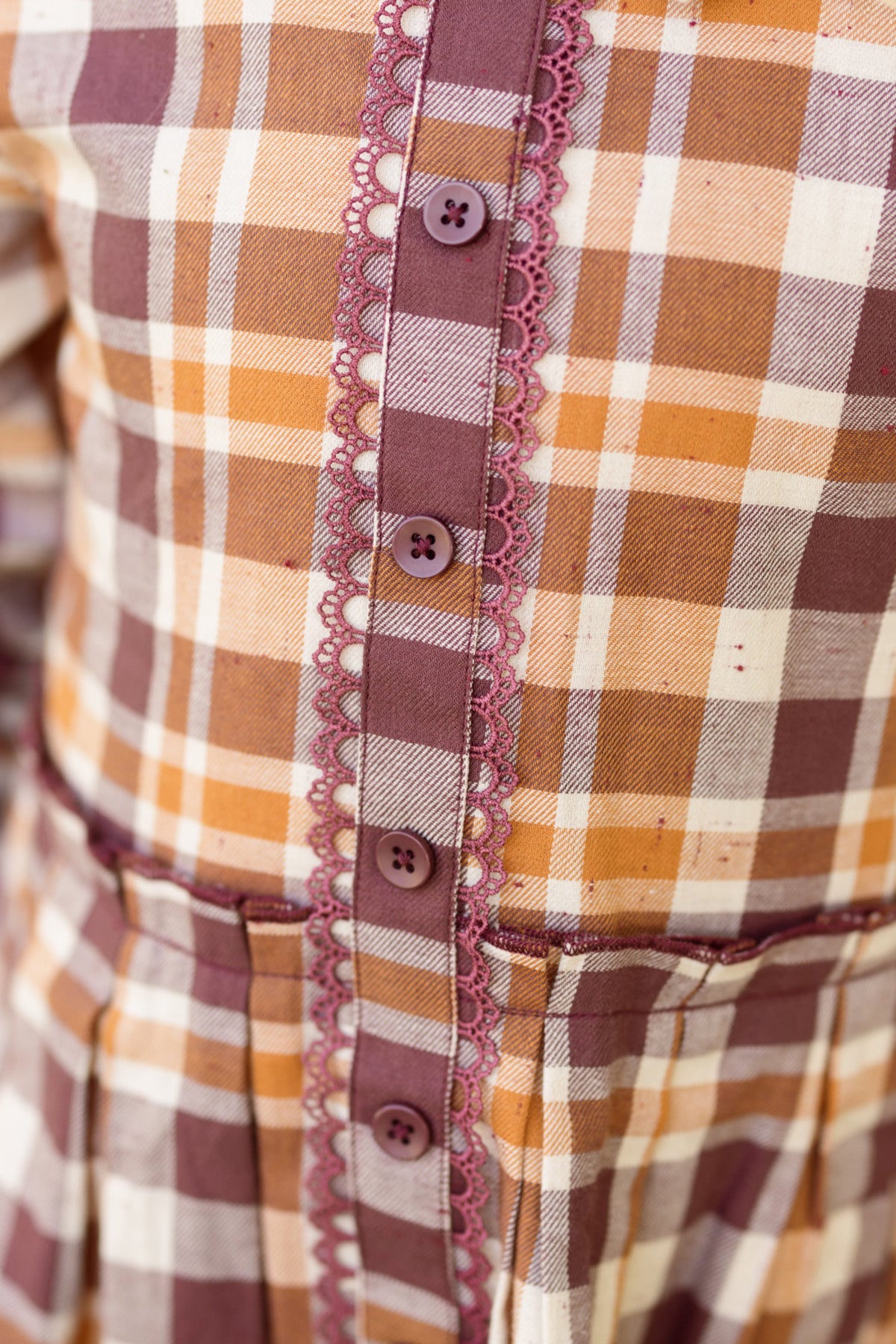 Close-up of the Ivy City Co Mini Bowie Long Sleeve Dress in Spiced Plaid, crafted from plaid cotton in brown, beige, and white hues, and featuring balloon sleeves, purple buttons, and lace trim along the front button placket.