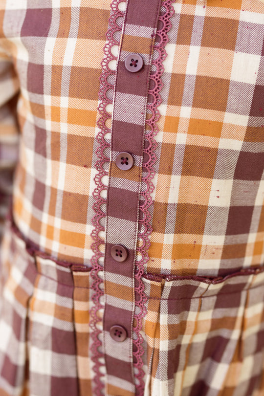 Close-up of the Ivy City Co Mini Bowie Long Sleeve Dress in Spiced Plaid, crafted from plaid cotton in brown, beige, and white hues, and featuring balloon sleeves, purple buttons, and lace trim along the front button placket.