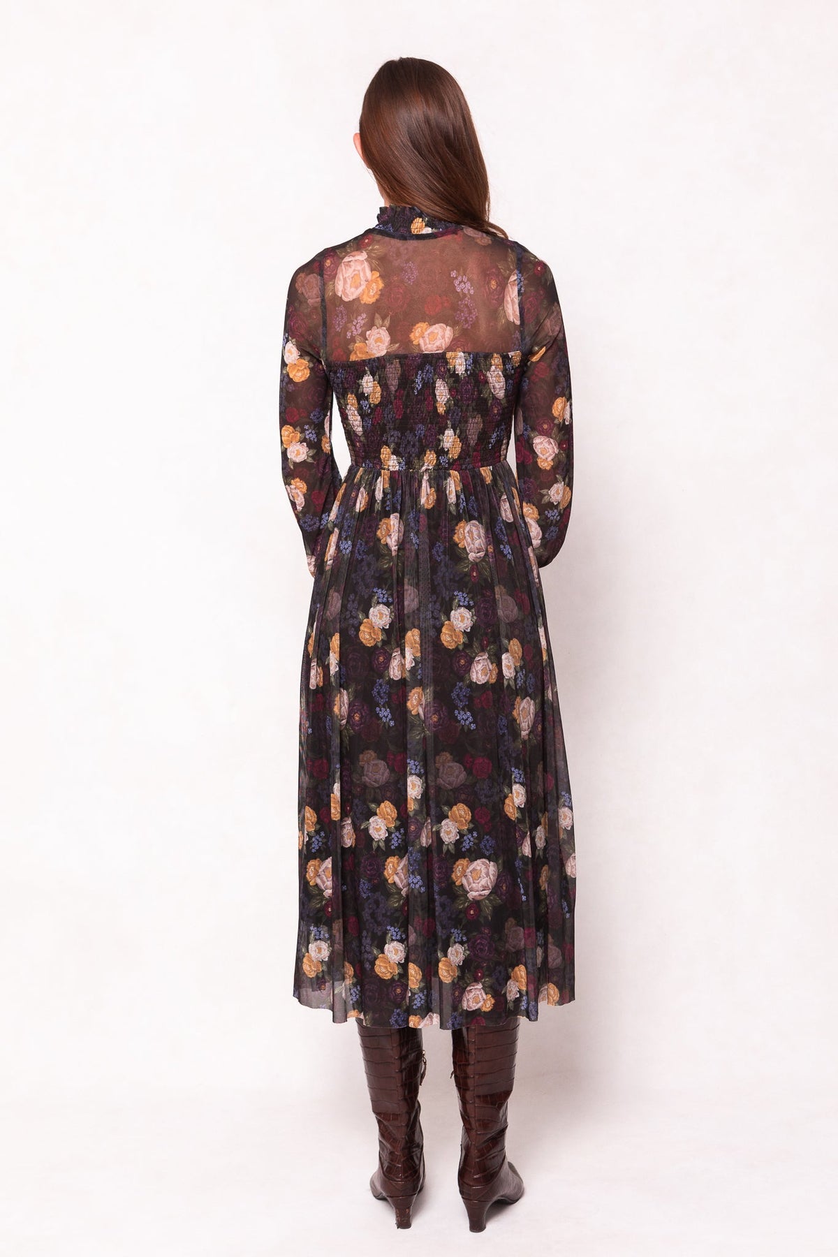 A person stands with their back to the camera, wearing the Ivy City Co Marla Dress in Winter Floral—a maternity-friendly, long-sleeve, sheer black floral dress—paired with knee-high brown boots.