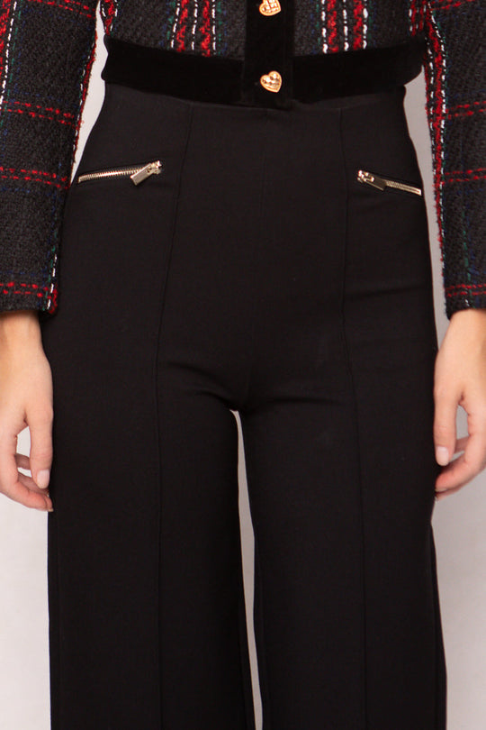 A person is wearing the Alyssa Pop Ponte Pants in Black by Ivy City Co, styled with a black, red, and green plaid jacket featuring gold zipper pockets—a perfect day-to-night staple. Only the torso and upper legs are visible.