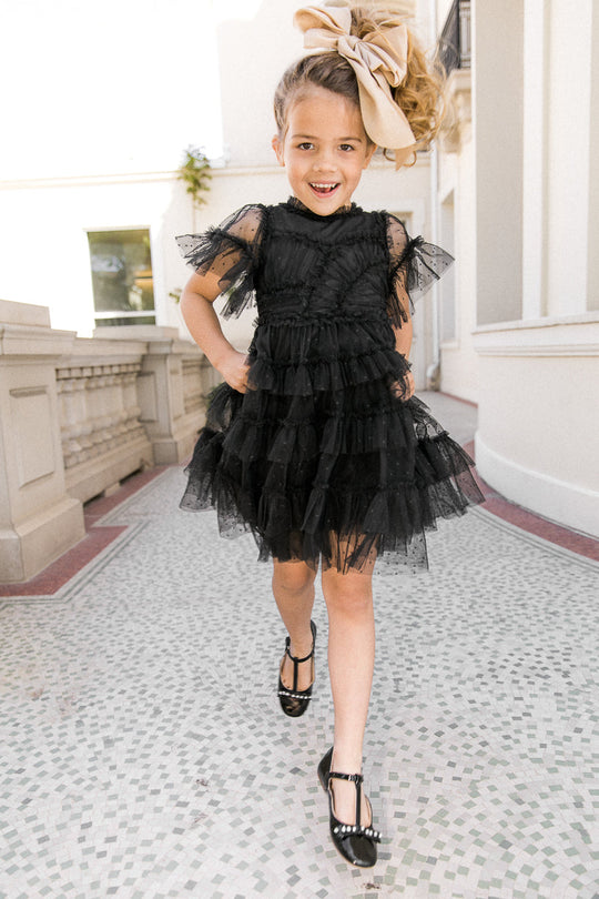 A smiling young girl wearing the Ivy City Co Mini Whimsical Dress in Black, paired with black shoes and a large beige hair bow, walks forward on a patterned stone path beside a white building.