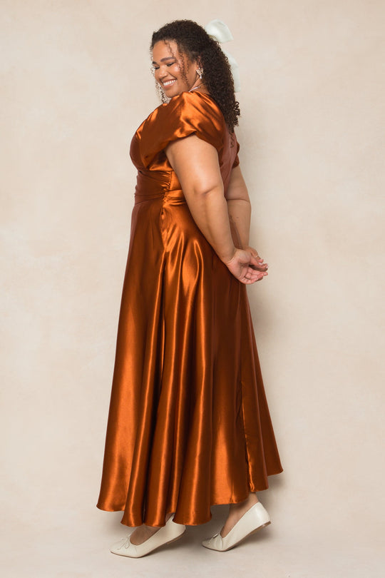 A woman wearing the Ivy City Co Tessie Dress in Amber Satin and white heels stands sideways, smiling with her hands clasped behind her back against a light beige background.