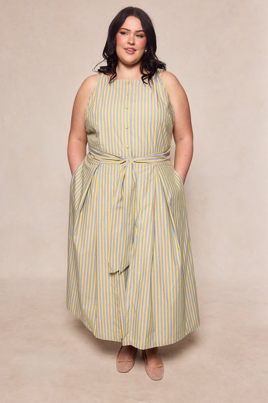 A woman stands wearing the Ivy City Co Glen Dress in Green Stripe, a sleeveless cotton ankle-length dress featuring green and white vertical stripes, buttons down the front, a waist tie, and pockets. She has dark wavy hair and beige flats, posing against a plain beige background.