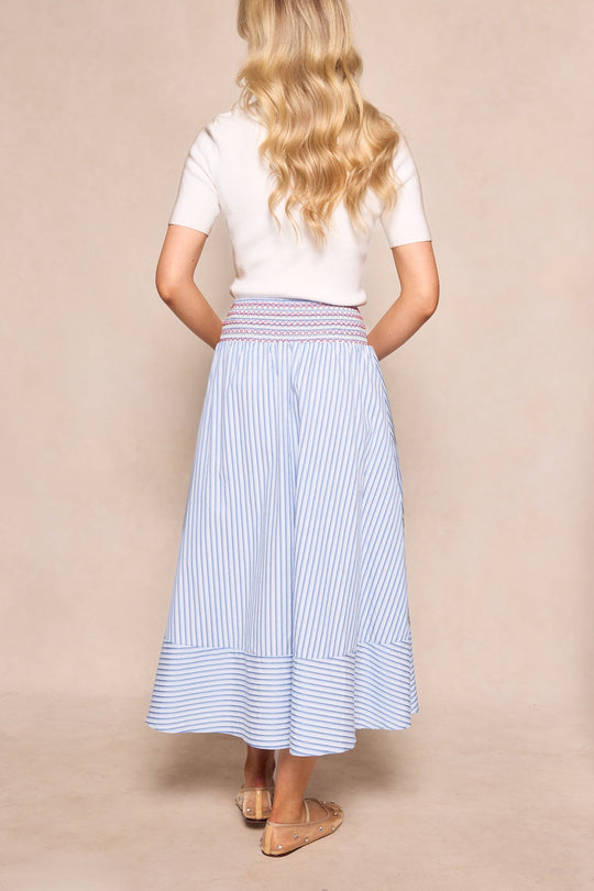 A woman with long blonde wavy hair stands with her back to the camera, wearing a white short-sleeve top, tan flat sandals, and the Ivy City Co Bluebell Skirt in Blue & White Stripes, which features a maternity-friendly smocked waistband.