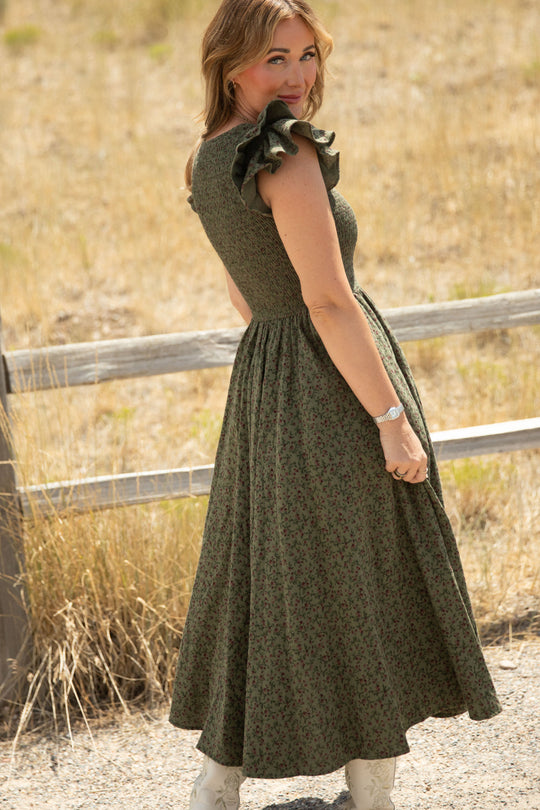 Dressed in the Ivy City Co Hattie Dress in Woodland Floral, this woman stands outdoors near a wooden fence, smiling over her shoulder on a sunny day with dry grass in the background; the flowy silhouette and smocked bodice of this ruffled-sleeve dress make it effortlessly maternity friendly.
