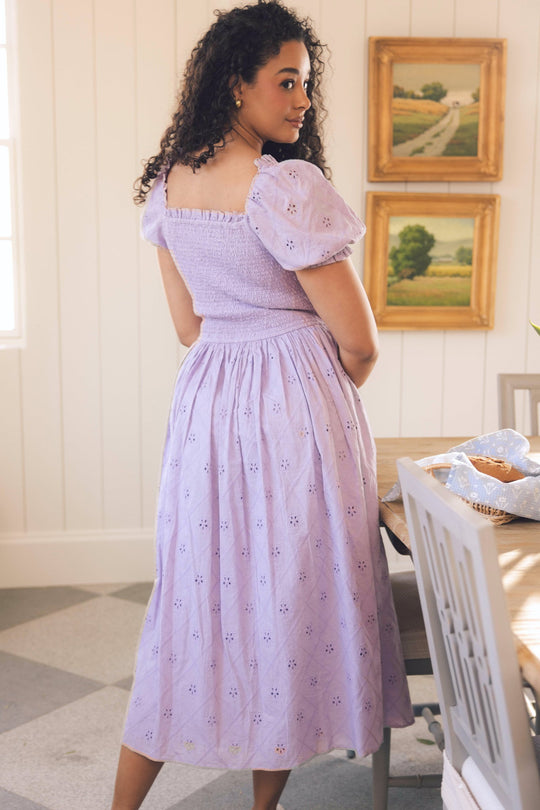 A woman with curly hair in the Emmi Dress in Lilac by Ivy City Co stands at a table, glancing over her shoulder. The room features light walls, framed artwork, and natural light streaming through a window.