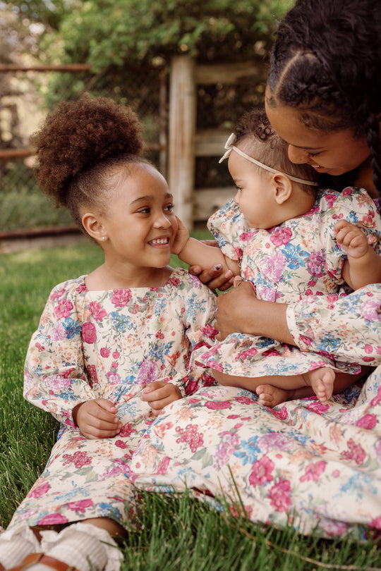 A woman holds a baby and sits next to a young girl, all three smiling at each other while wearing matching Ivy City Co. Mini Adelpha Dresses in Autumn Amity Floral, featuring smocked bodices and balloon sleeves as they sit together on the grass outdoors.