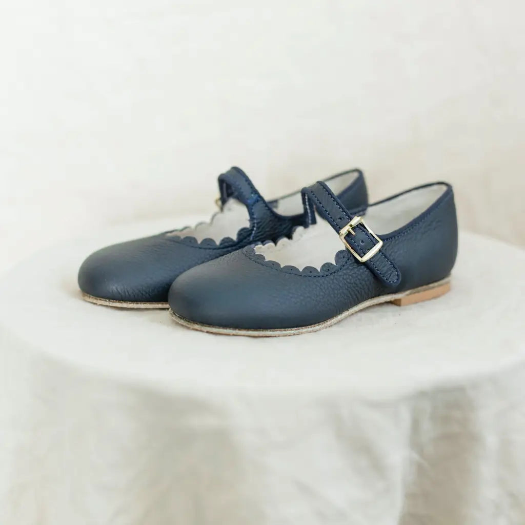 A pair of Mini Mary Janes in Scalloped Navy by Zimmerman Shoes, featuring scalloped edges and buckle straps, are displayed on a round, white, textured surface.