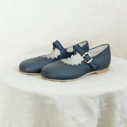 A pair of Mini Mary Janes in Scalloped Navy by Zimmerman Shoes, featuring scalloped edges and buckle straps, are displayed on a round, white, textured surface.