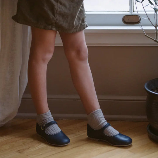 A child wearing a brown skirt, gray socks, and Zimmerman Shoes’ Mini Mary Janes in Scalloped Navy stands on a wooden floor near a window and a plant pot.
