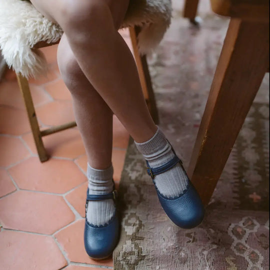 A person with crossed legs sits on a chair, wearing gray socks and the Mini Mary Janes in Scalloped Navy by Zimmerman Shoes, featuring an adjustable ankle strap, placed on a patterned rug and terracotta tile floor.
