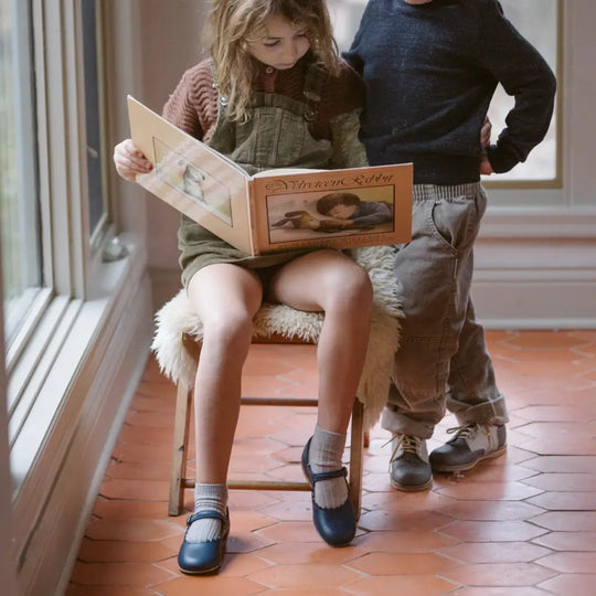 A child wearing Zimmerman Shoes’ Mini Mary Janes in Scalloped Navy with an adjustable ankle strap sits on a stool reading a picture book, while another child stands nearby. They are indoors on a terracotta tiled floor near a window.