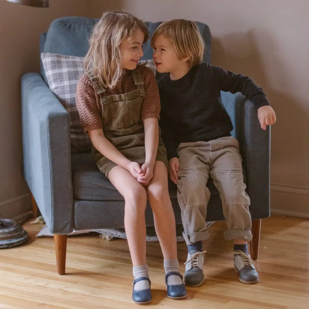 Two young children sit closely together on an armchair, smiling and looking at each other. The girl is wearing overalls, a brown top, and the Mini Mary Janes in Scalloped Navy by Zimmerman Shoes—which feature a classic scalloped edge design and an adjustable ankle strap—as she shares a warm moment with the boy in a dark sweater and light pants. They appear happy and relaxed in a cozy room.