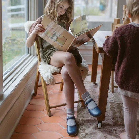 A young girl sits by a window on a wooden chair, reading a book. She wears a green dress, blue socks, and the Mini Mary Janes in Scalloped Navy by Zimmerman Shoes, featuring an adjustable ankle strap and classic navy leather. Another child stands nearby at a table with wooden blocks as natural light fills the cozy room.
