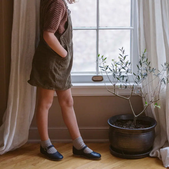 A child in a brown shirt and overalls stands by a window with hands in pockets, next to a small potted plant on a wooden floor, wearing Zimmerman Shoes’ Mini Mary Janes in Scalloped Navy.