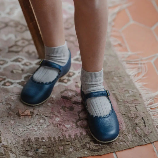 A child wearing white socks and Zimmerman Shoes’ Mini Mary Janes in Scalloped Navy with an adjustable ankle strap stands on a patterned rug near a tiled floor.