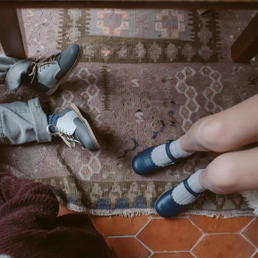 Two people sit at a table on a patterned rug, their legs and shoes visible. One wears gray pants with boots, while the other sports Zimmerman Shoes’ Mini Mary Janes in Scalloped Navy, styled with socks and featuring an adjustable ankle strap. The floor is terracotta tiles.