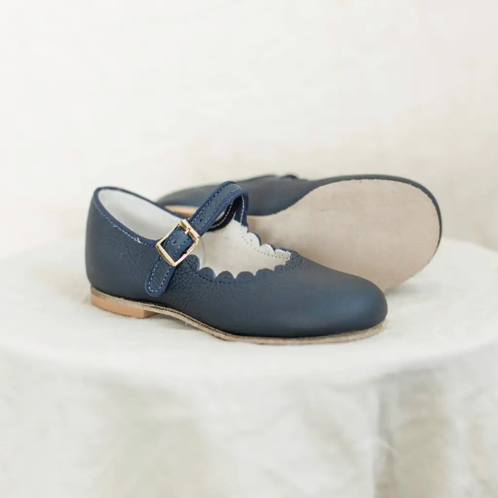 The Mini Mary Janes in Scalloped Navy by Zimmerman Shoes are children’s navy leather Mary Jane shoes with scalloped edges, gold buckles, light brown soles, and an adjustable ankle strap, displayed on a white fabric surface.