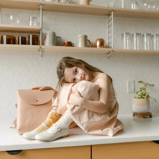 A young girl in a light dress sits on a kitchen counter, hugging her knees beside a pink bag. She wears Zimmerman Shoes’ Mini Mary Janes in Scalloped Pearl with an adjustable ankle strap. Shelves lined with jars, cups, and a potted plant are visible in the background.