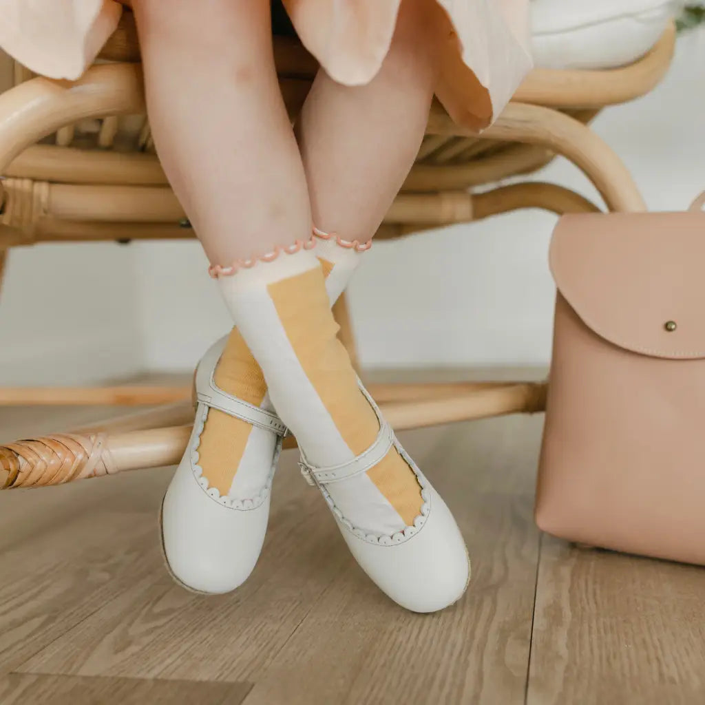 A child sits on a wicker chair with legs crossed, wearing Zimmerman Shoes’ Mini Mary Janes in Scalloped Pearl—white leather shoes featuring an adjustable ankle strap—paired with yellow-and-white socks. A light pink bag rests on the wooden floor beside them.